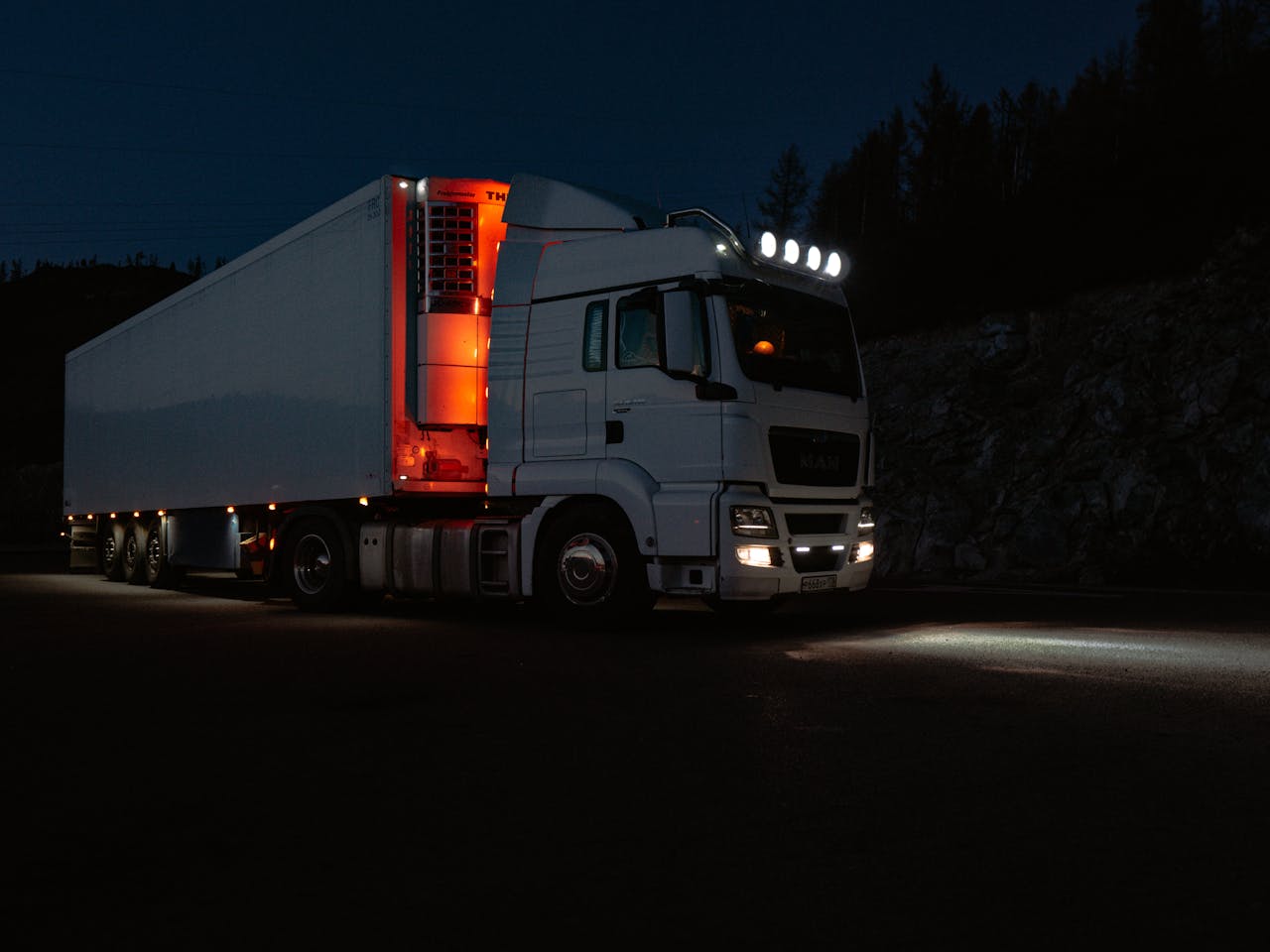A white truck lit by headlights and interior lights on a dark road in mountainous terrain.